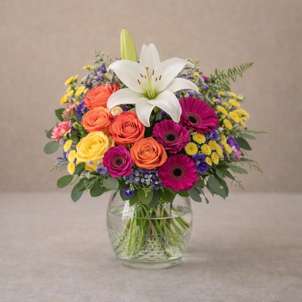 Premium size Radiant Cheer bouquet showcasing a white lily, colorful gerberas and roses with a lush, expanded silhouette in a clear hourglass glass vase — Encinitas flower delivery.