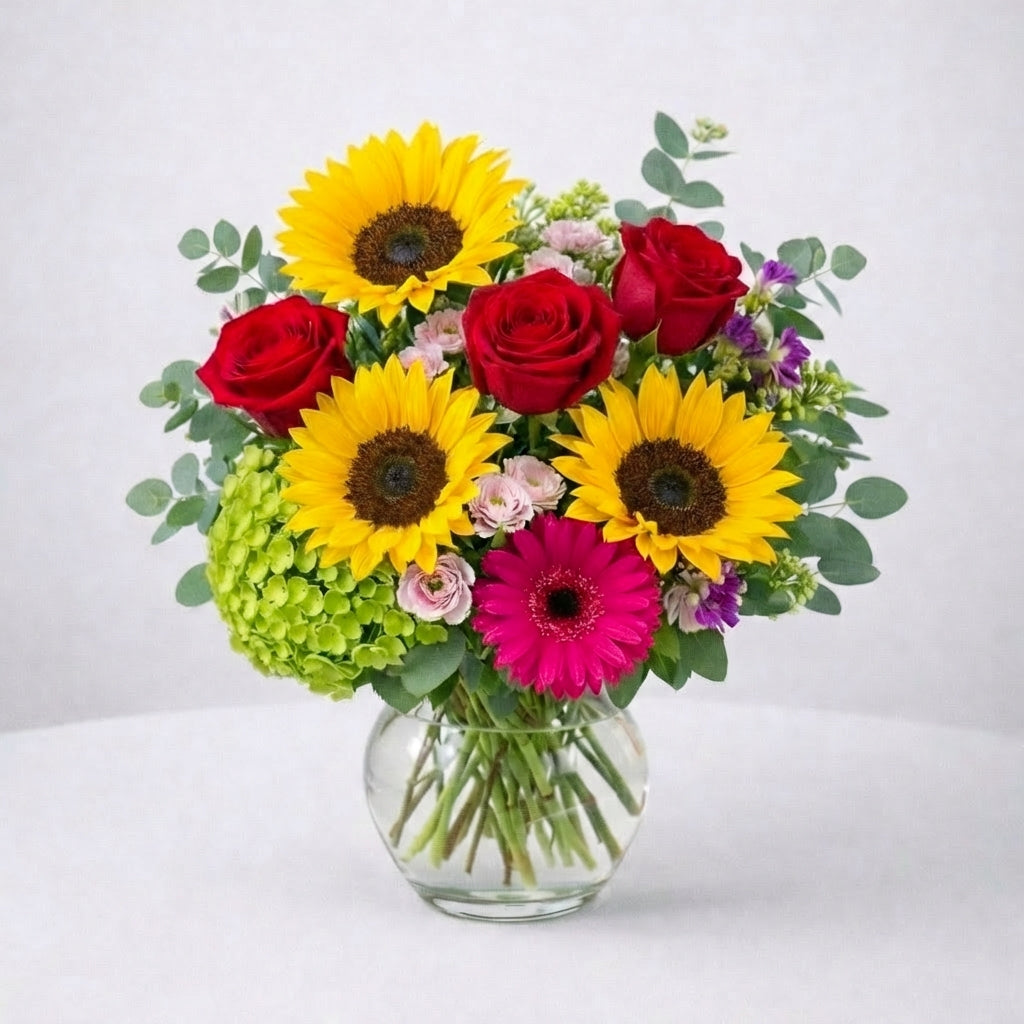 Lush premium sunflower and red rose arrangement with abundant pink gerbera daisies, green accents, and a full rounded design in a clear glass vase β same-day flower delivery Oceanside.