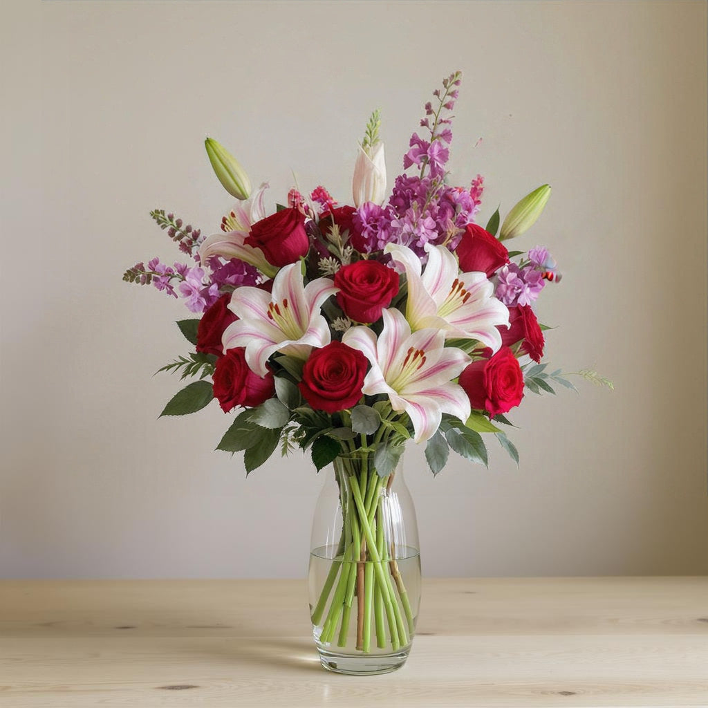 Premium size luxurious red rose and pink lily bouquet featuring abundant blooms, vivid purple accents, and rich greenery arranged in a clear glass vase on a softly lit background — North County San Diego flower delivery.