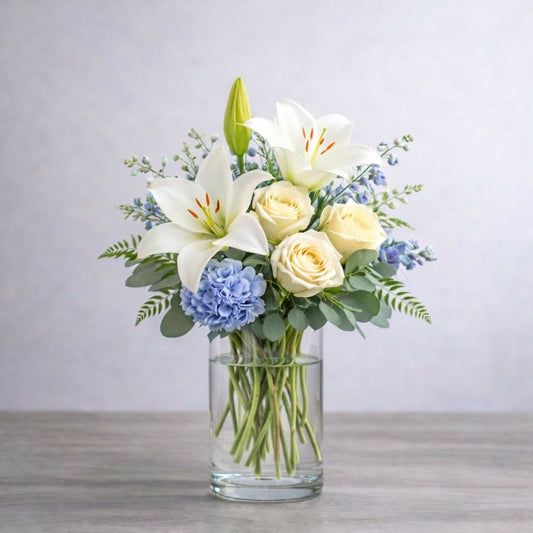 Regular size white lily and cream rose vase arrangement with blue hydrangea, soft delphinium, baby’s breath, and fresh greenery in a clear glass vase. Fresh flowers San Marcos CA.