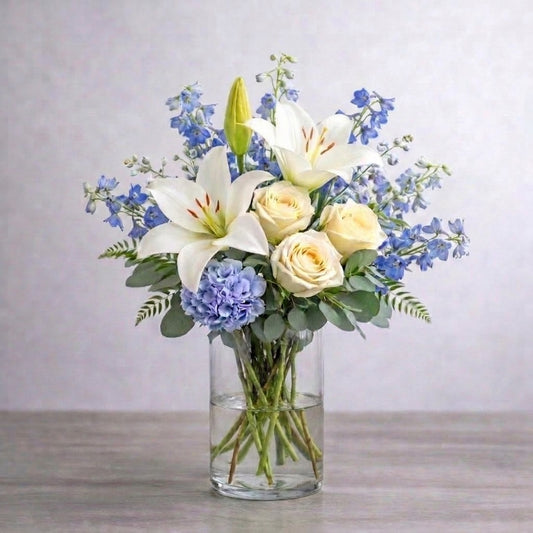 Regular size white lily and cream rose vase arrangement featuring fuller blue hydrangea, layered delphinium, baby’s breath, and lush greenery for a calm, elegant look. Fresh flowers San Marcos CA.