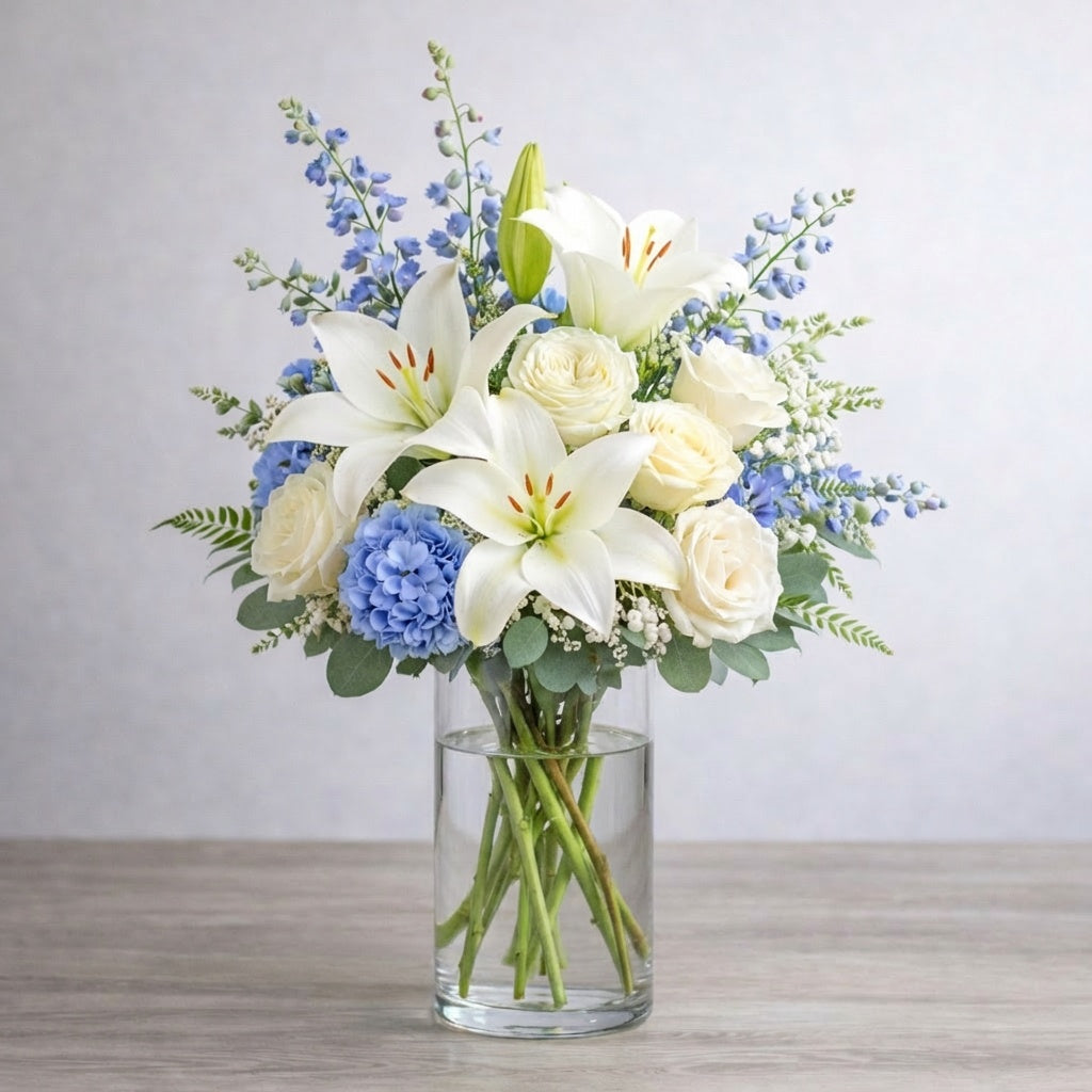 Full size white lily and cream rose vase arrangement featuring fuller blue hydrangea, layered delphinium, baby’s breath, and lush greenery for a calm, elegant look. Fresh flowers San Marcos CA.