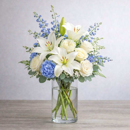 Full size white lily and cream rose vase arrangement featuring fuller blue hydrangea, layered delphinium, baby’s breath, and lush greenery for a calm, elegant look. Fresh flowers San Marcos CA.