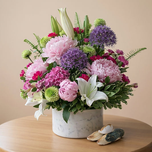 Elegant mixed flower arrangement featuring pink peonies, purple alliums, white lilies, and lush greenery in a white marble vase on a wooden table — San Marcos flower delivery.