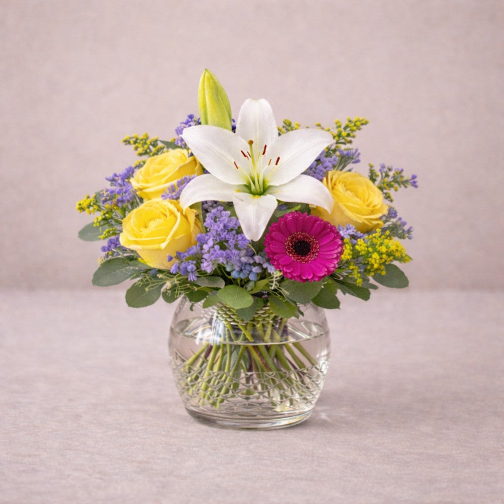 Regular size Radiant Cheer bouquet with white lily, pink gerbera daisies, yellow and orange roses arranged in a clear hourglass glass vase — Encinitas flower delivery.