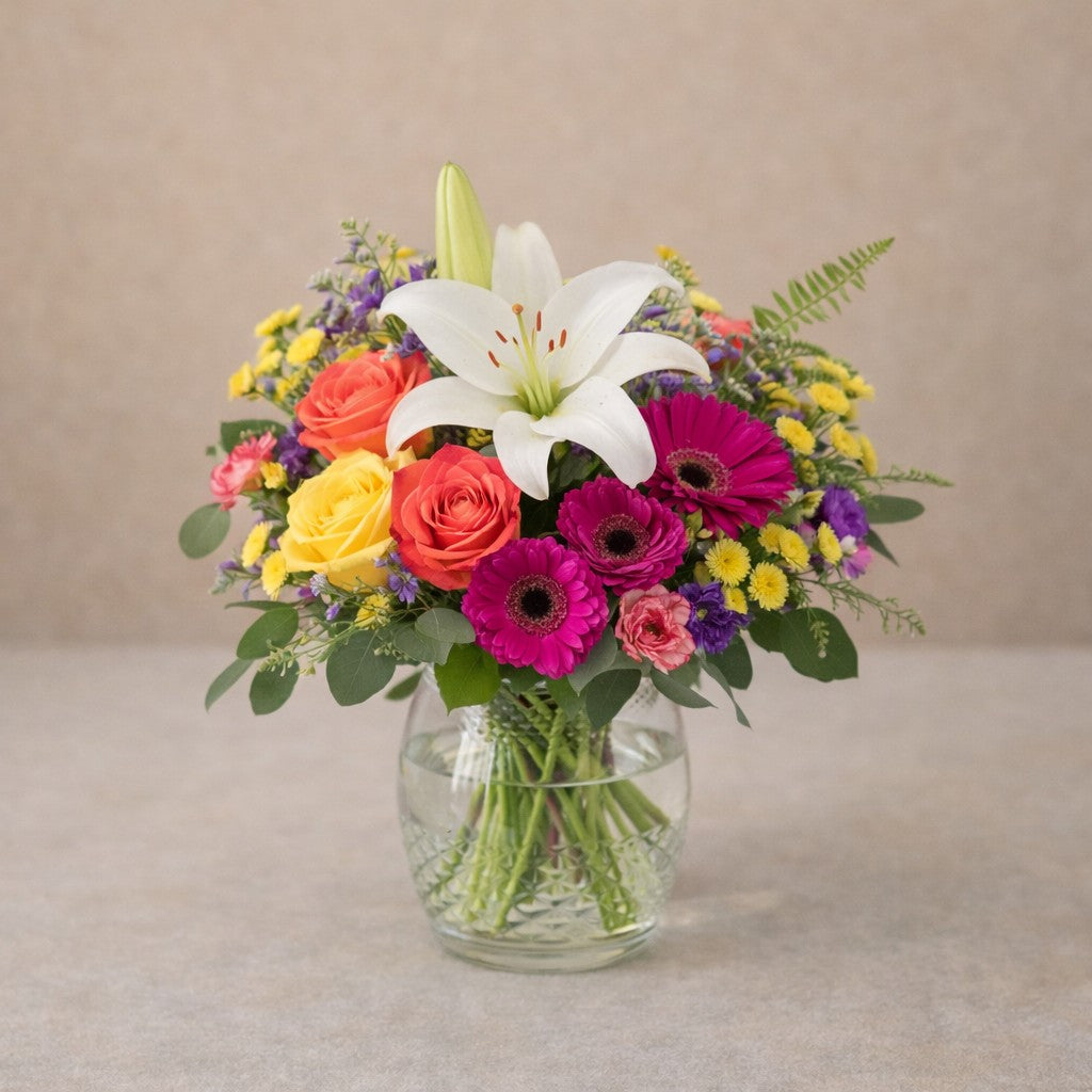 Deluxe size Radiant Cheer bouquet featuring a white lily, vibrant gerberas, yellow and orange roses with fuller volume in a clear hourglass glass vase — Encinitas flower delivery.