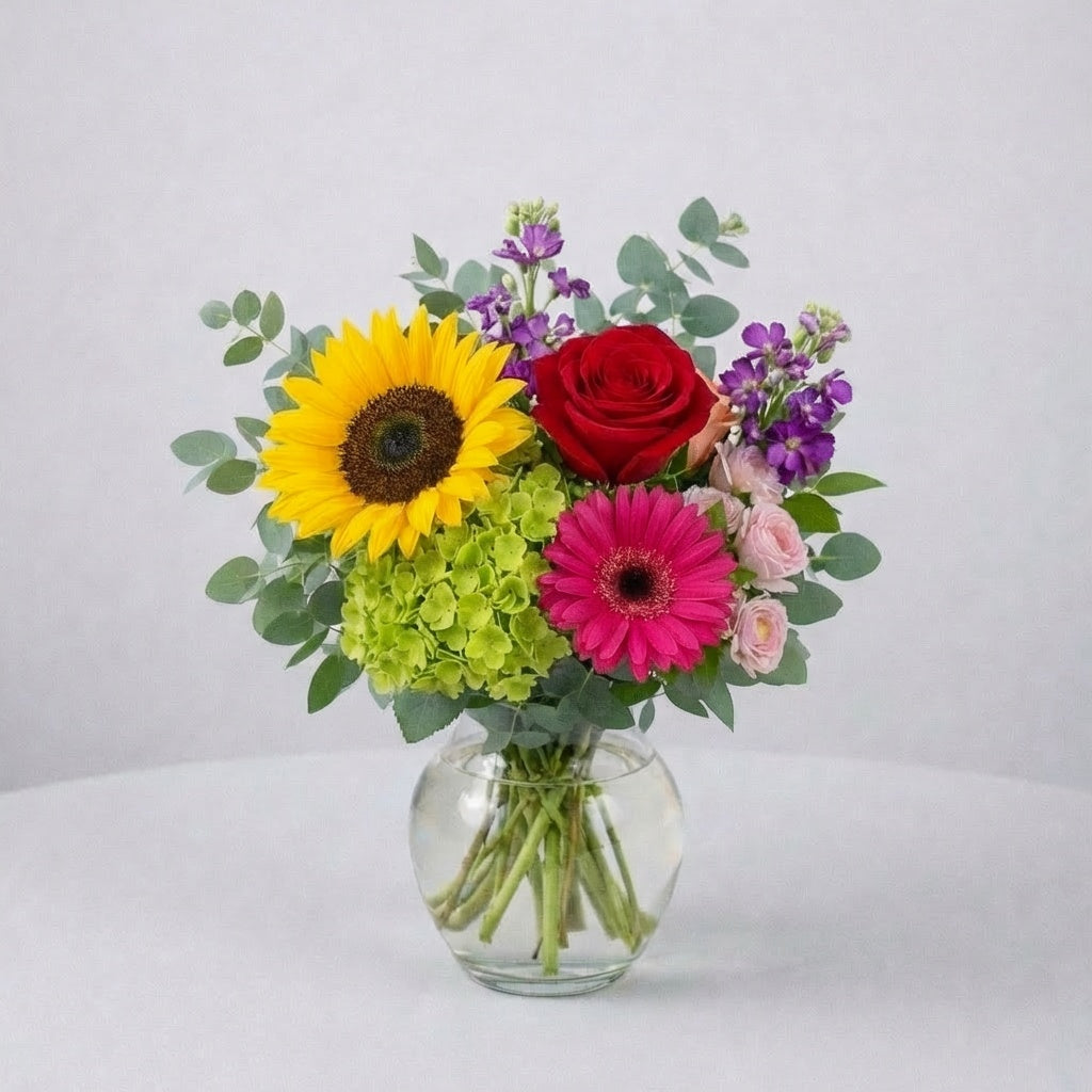Bright sunflower and red rose vase arrangement with pink gerbera daisies and fresh greenery in a clear round glass vase — same-day flower delivery Oceanside.