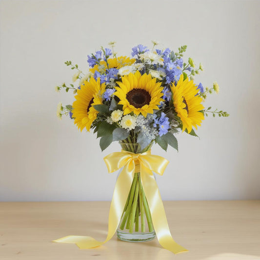 Regular size sunflower and blue aster flower arrangement featuring bright sunflowers, soft blue accent blooms, yellow filler flowers, and eucalyptus greenery tied with a yellow ribbon in a clear glass vase — Local florist in North San Diego County.