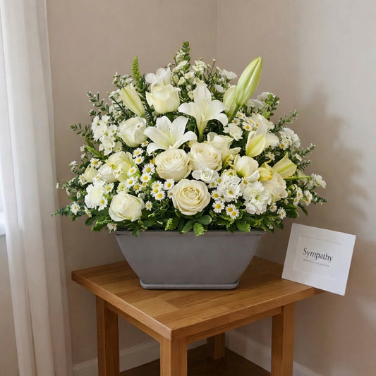 Elegant sympathy basket arrangement with white roses, lilies, and soft yellow blooms in a modern gray container - Vista florist.
