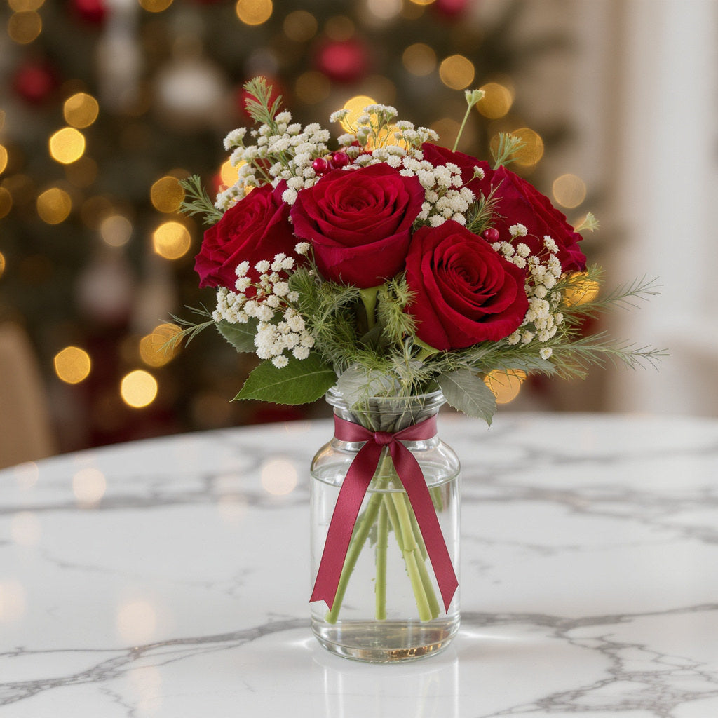 Regular size holiday rose bouquet arranged with red roses, white filler flowers, greenery, and a pink ribbon in a clear vase – San Marcos florist.