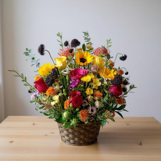Vibrant mixed flower arrangement in a woven basket featuring sunflowers, red roses, and assorted seasonal blooms on a wooden table - Oceanside flower delivery.