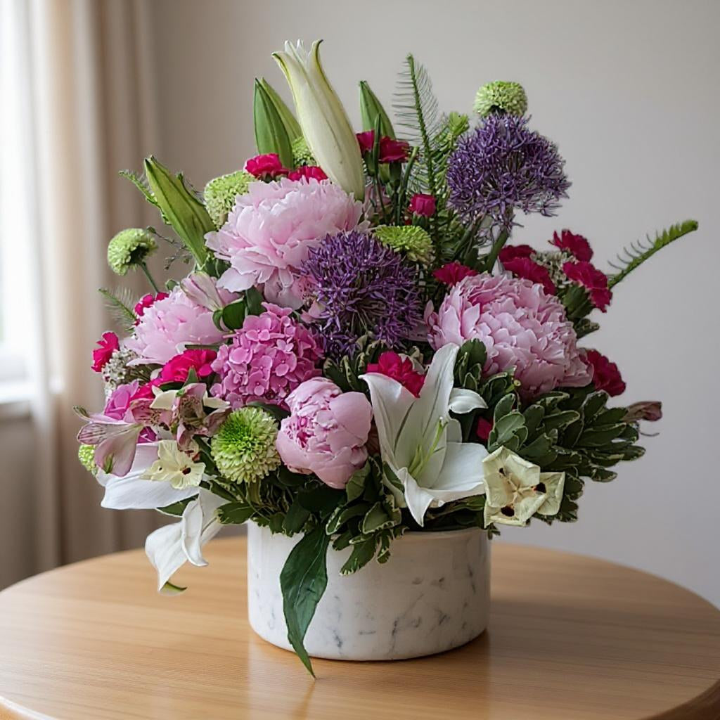 Elegant mixed flower arrangement featuring pink peonies, purple alliums, white lilies, and lush greenery in a white marble vase on a wooden table — San Marcos flower delivery.