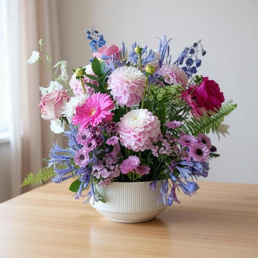 Pastel pink and lavender flower arrangement in a white bowl-shaped vase on a wooden table, featuring roses, chrysanthemums, and delphiniums - Escondido florist.