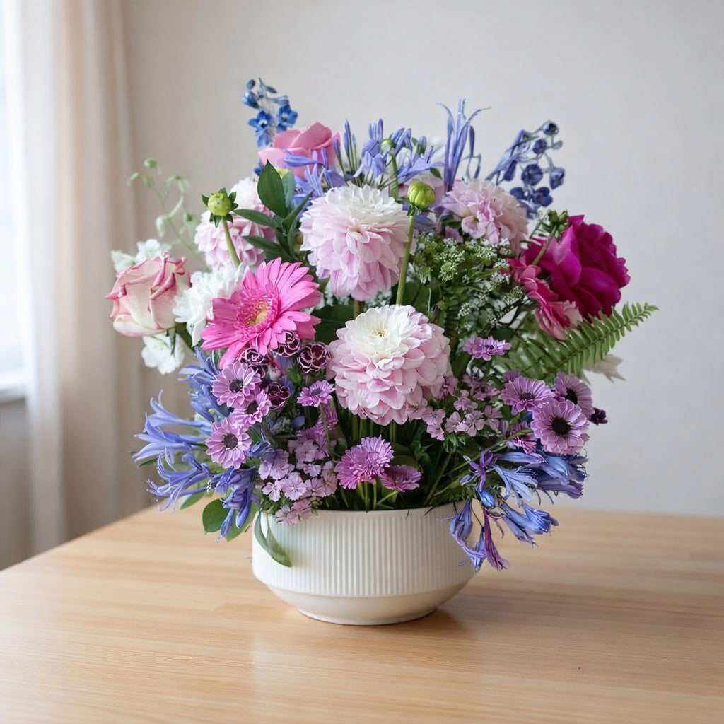 Pastel pink and lavender flower arrangement in a white bowl-shaped vase on a wooden table, featuring roses, chrysanthemums, and delphiniums - Escondido florist.