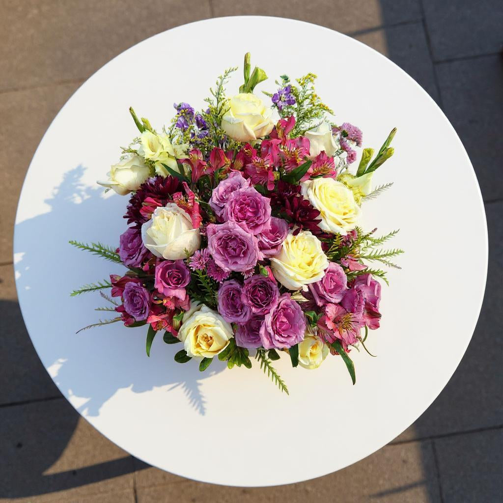 Top view of a mixed rose basket arrangement featuring purple roses, cream roses, greenery, and filler flowers in a woven wicker basket - Carlsbad flower delivery.