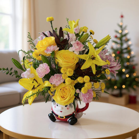 Yellow and pink Christmas flower arrangement with roses, lilies, carnations, and button mums in a Santa mug vase on a white table in a cozy holiday living room – North San Diego County flower delivery.