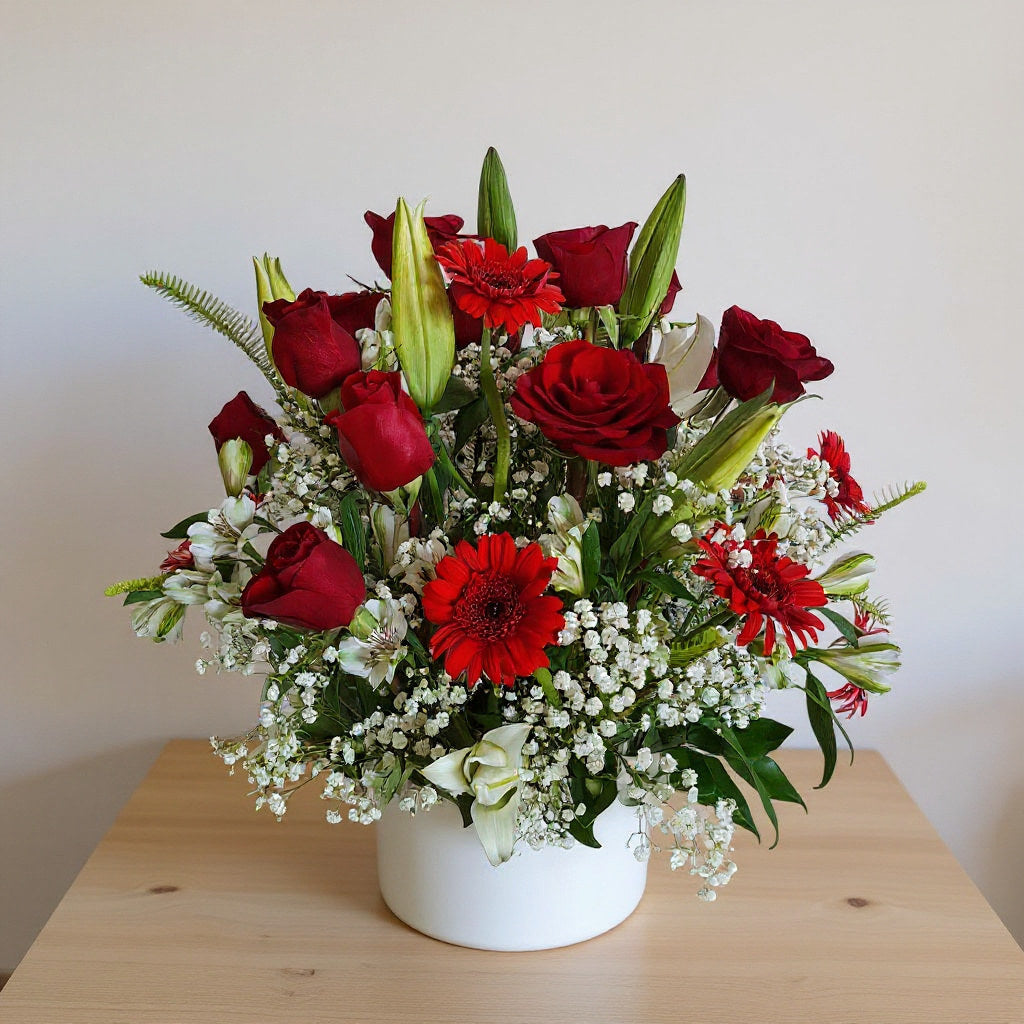 Red roses, red gerbera daisies, white baby's breath, and unopened lilies arranged in a white ceramic vase on a wooden table - Vista florist.