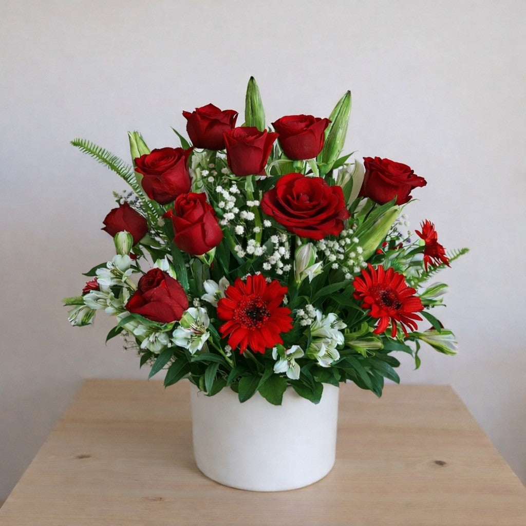 Red roses, red gerbera daisies, white baby's breath, and unopened lilies arranged in a white ceramic vase on a wooden table - Vista florist.