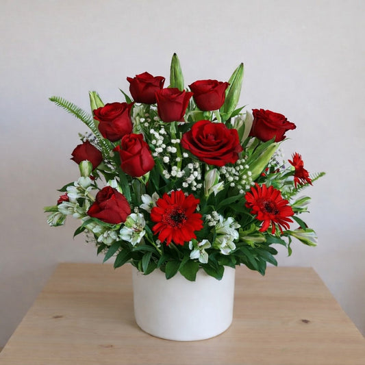 Red roses, red gerbera daisies, white baby's breath, and unopened lilies arranged in a white ceramic vase on a wooden table - Vista florist.