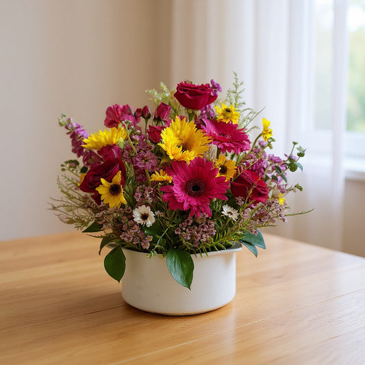 Bright mixed flower arrangement with pink gerbera daisies, yellow blooms, red ranunculus, and wildflower accents in a white vase on a wooden table in natural window light - Same-day flowers San Marcos.