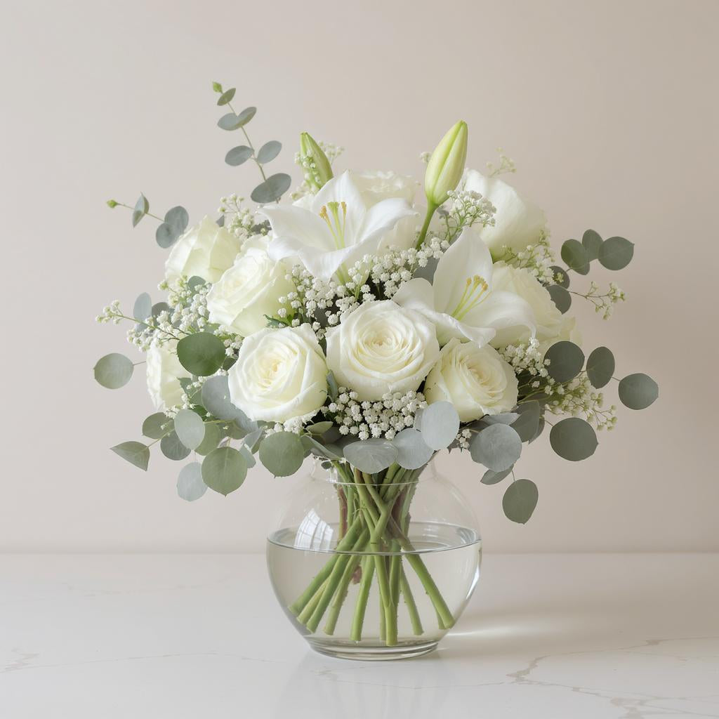 Deluxe size elegant white flower arrangement featuring white lilies, cream roses, baby’s breath, and eucalyptus greenery arranged in a round clear glass vase — Carlsbad florist.