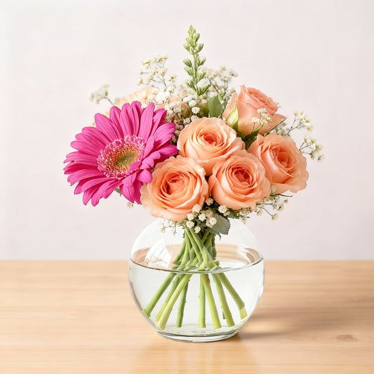 Regular size bouquet with bright pink gerbera daisies, peach roses, white fillers, and eucalyptus arranged in a round bowl glass vase on a wooden table — North San Diego County florist.