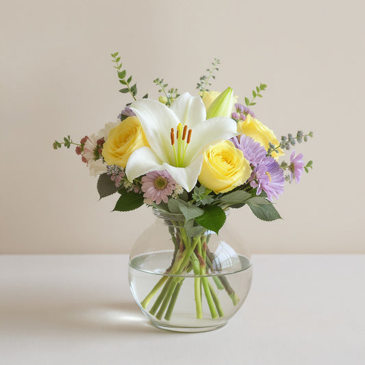 Regular size white lily and yellow rose bouquet with pastel accents and greenery in a clear round vase — Escondido flower delivery.