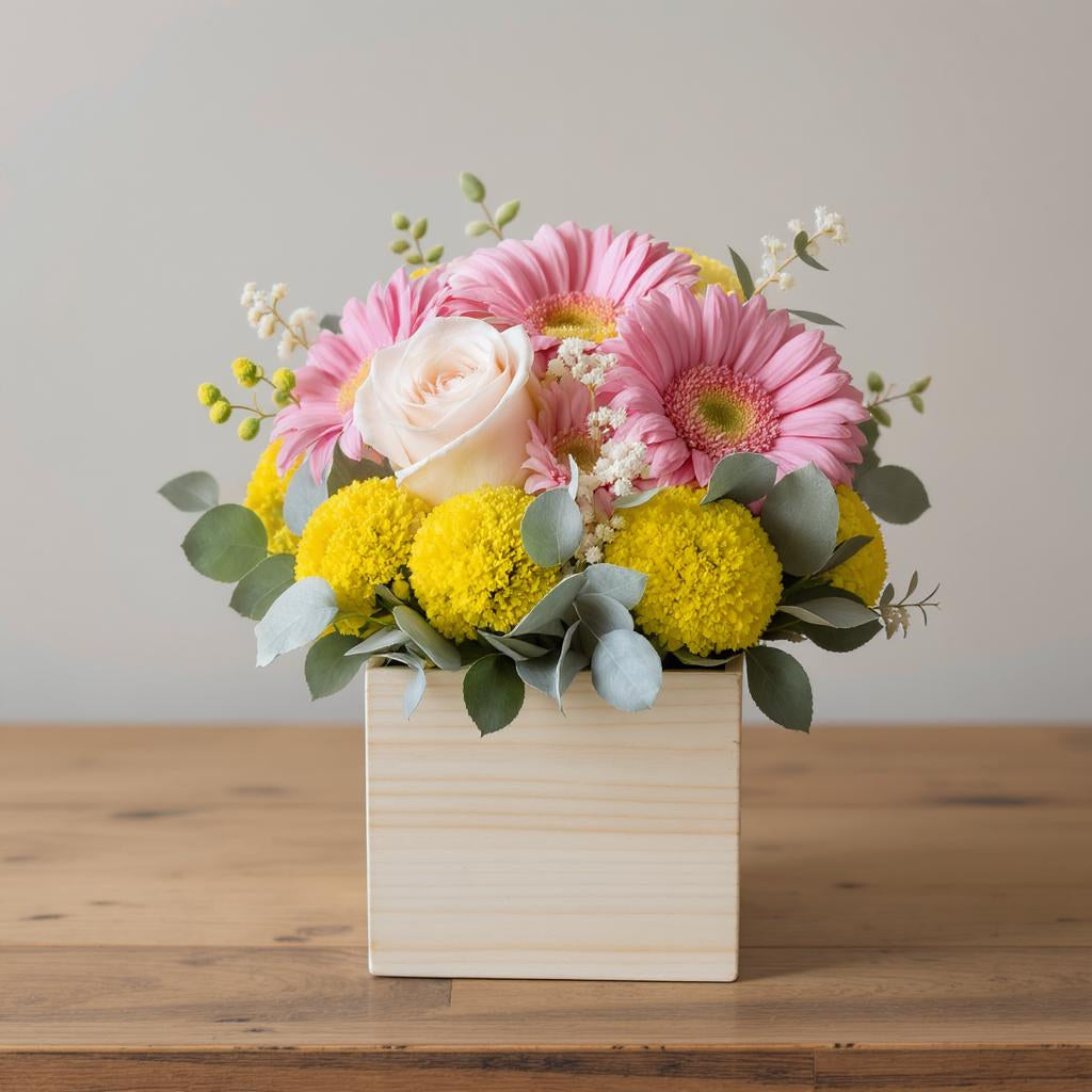 Full size cheerful flower arrangement featuring pink gerberas, pale blush roses, yellow billy balls, and eucalyptus greenery arranged in a light wooden square container — Same-day flowers Carlsbad.