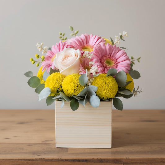 Regular size cheerful flower arrangement featuring pink gerberas, pale blush roses, yellow billy balls, and eucalyptus greenery arranged in a light wooden square container — Same-day flowers Carlsbad.