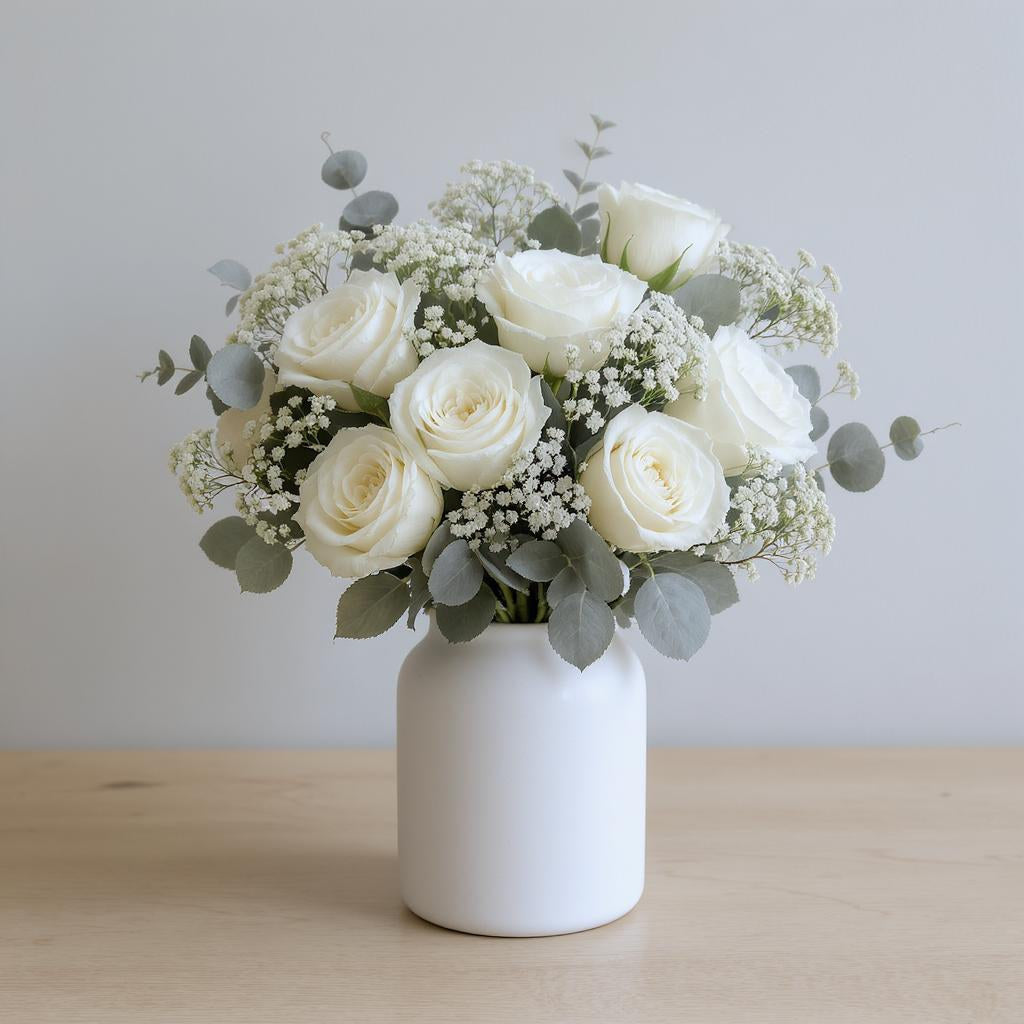 Deluxe size white rose arrangement featuring cream roses, baby’s breath, eucalyptus, and mixed greenery in a tall white ceramic vase with a warm indoor background - Escondido flower delivery.