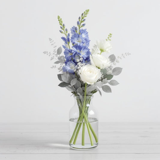 Regular size elegant flower arrangement with blue delphiniums, white roses, white ranunculus, and silver eucalyptus in a tall clear glass vase on a light wooden surface - Vista florist.