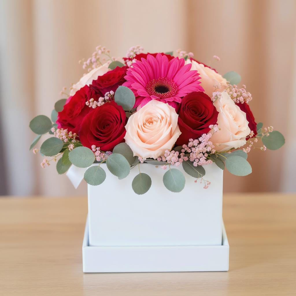 Regular size flower arrangement with pink gerbera daisies, red roses, blush roses, and eucalyptus in a white box vase on a wooden table - Oceanside florist.