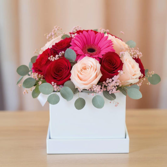 Regular size flower arrangement with pink gerbera daisies, red roses, blush roses, and eucalyptus in a white box vase on a wooden table - Oceanside florist.