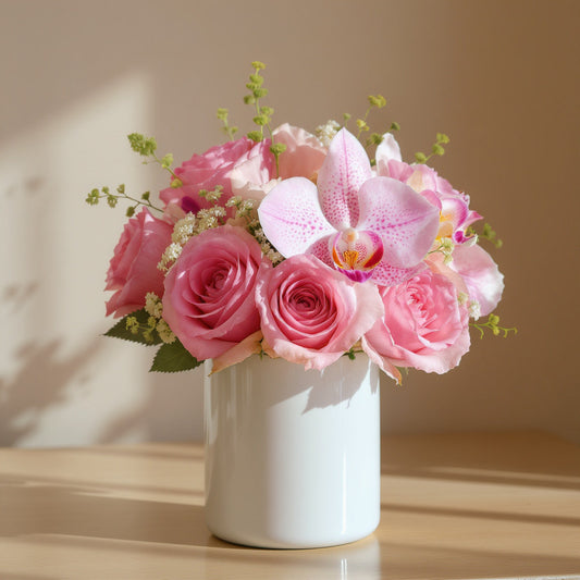 Regular size pink flower arrangement with pink roses, pink orchids, and soft greenery in a white ceramic vase on a wooden table in natural sunlight - Same-day flowers Vista.