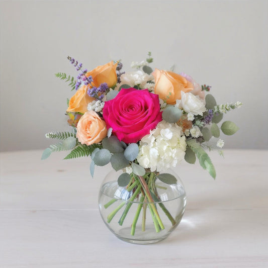 Regular size colorful flower arrangement featuring bright pink ranunculus, peach roses, white hydrangea, lavender accents, and eucalyptus greenery arranged in a round clear glass vase — Same-day flowers San Marcos.