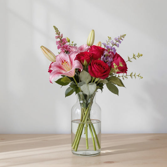 Regular size mixed flower arrangement featuring bright red roses, pink lilies, purple accent blooms, and greenery arranged in a clear glass vase on a wooden surface — North County San Diego flower delivery.